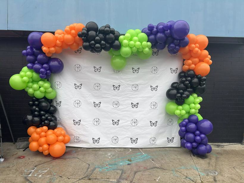 Custom balloon arch in orange, green, purple and black framing a printed photo backdrop