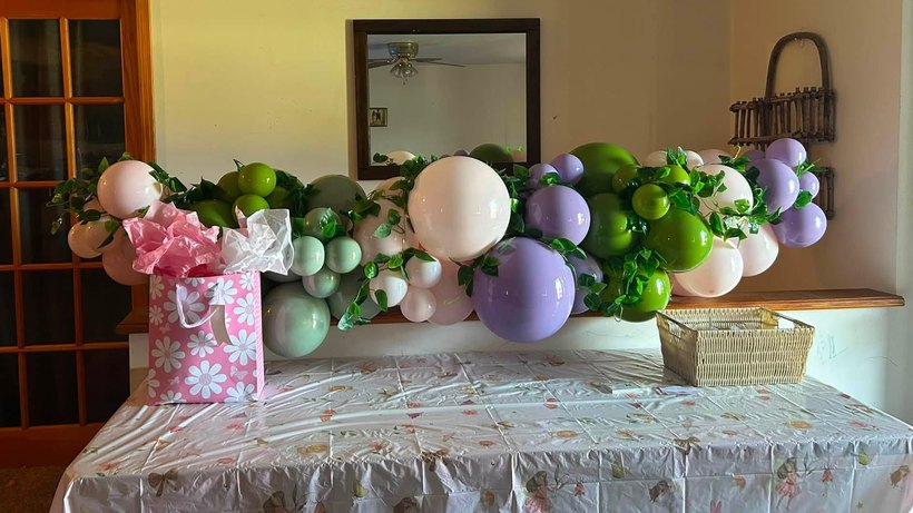 Balloon garland in sage green, olive, lavender and white threaded with ivy running along a ledge above a styled gift table