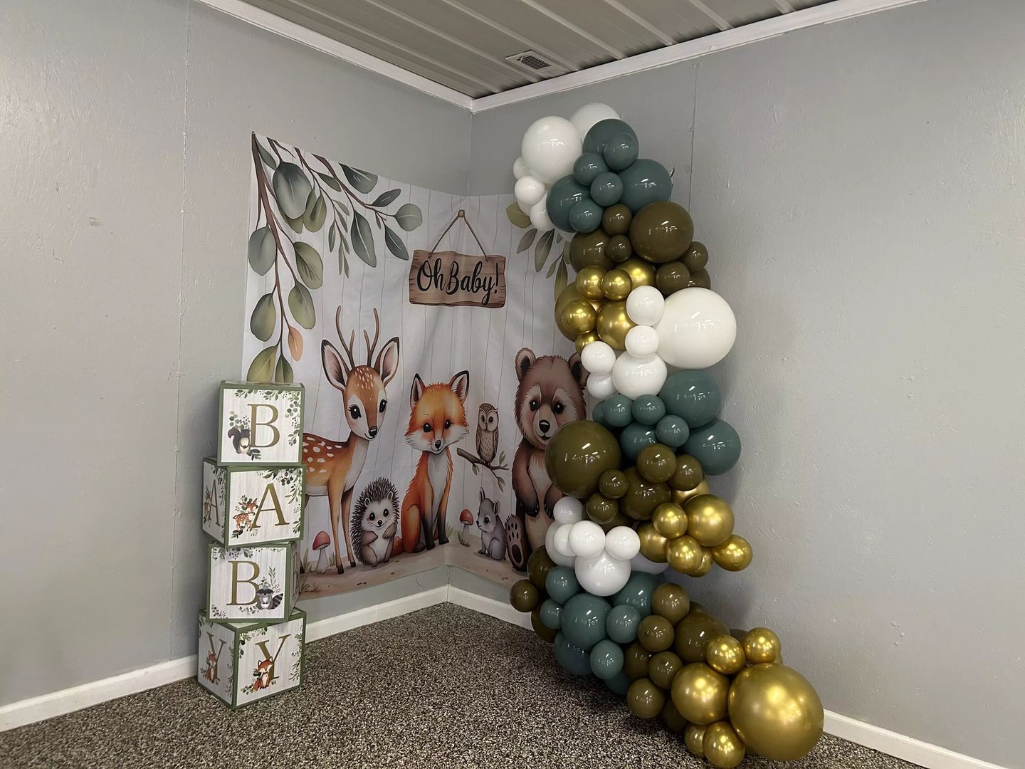 Woodland baby shower balloon garland in sage green, gold and white curving down beside an Oh Baby backdrop and stacked letter blocks